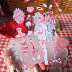 Puede incluir: Una hoja de pegatinas transparente con ilustraciones m&eacute;dicas en tonos rosas que incluyen un personaje de enfermer&iacute;a, un soporte para gotero, una jeringa grande, un coraz&oacute;n, dientes, pastillas y alas. El texto "nisestarmin" aparece impreso.
