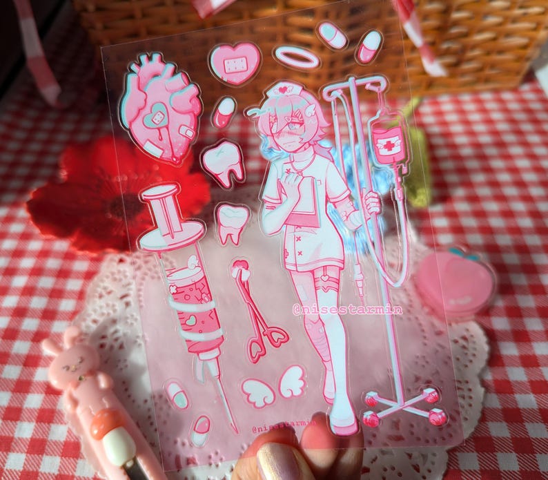 Puede incluir: Una hoja de pegatinas transparente con ilustraciones m&eacute;dicas en tonos rosas que incluyen un personaje de enfermer&iacute;a, un soporte para gotero, una jeringa grande, un coraz&oacute;n, dientes, pastillas y alas. El texto "nisestarmin" aparece impreso.