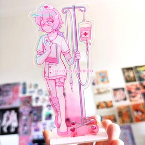 Puede incluir: Una figura de acrílico con un personaje de estilo anime en tonos rosas vestido de enfermero, sosteniendo un portapapeles y junto a un soporte de suero con una bolsa. El texto @nisestarmin está impreso en la figura sobre una base cuadrada.