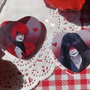 Puede incluir: Dos pines en forma de corazón con ilustraciones de estilo anime. Uno presenta un personaje pelirrojo con abrigo rojo y paraguas. El otro muestra un personaje con pelo largo y negro. Ambos pines tienen un corazón.