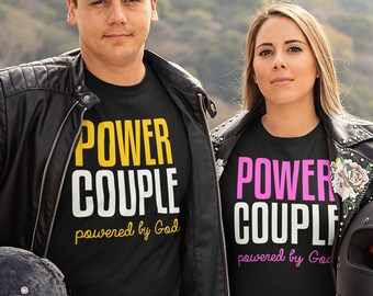 Könnte beinhalten: Ein Paar trägt schwarze T-Shirts mit dem Text "POWER COUPLE powered by God" in gelber und rosa Schrift. Der Mann trägt eine schwarze Lederjacke und die Frau trägt eine schwarze Lederjacke mit silbernen Nieten. Sie stehen vor einem Motorrad.