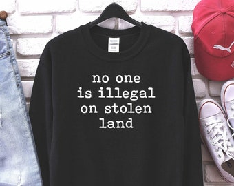 Könnte beinhalten: Schwarzer Pullover mit dem Text "no one is illegal on stolen land" in weißen Buchstaben. Der Pullover ist an einer weißen Backsteinwand mit einer blauen Jeans, einer roten Baseballkappe und weißen Turnschuhen ausgestellt.