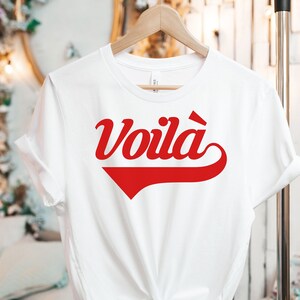 Puede incluir: Camiseta blanca con un gr&aacute;fico rojo "Voila!" en una fuente de b&eacute;isbol retro.