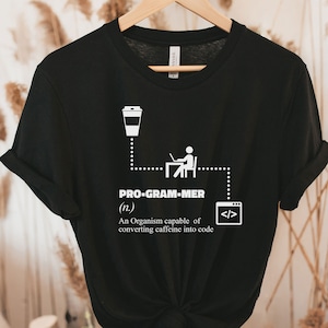 Puede incluir: Camiseta negra con un diseño gráfico blanco que define a un programador como un organismo que convierte la cafeína en código. El gráfico incluye una taza de café, una persona sentada frente a un ordenador y símbolos de código.
