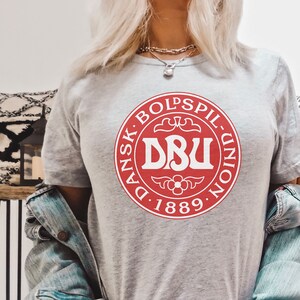 Puede incluir: Una camiseta gris con un logotipo circular rojo que dice "Dansk Boldspil-Union DBU 1889".