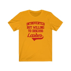 May include: Yellow short-sleeved t-shirt with red text that reads "Introverted but willing to discuss Lashes".