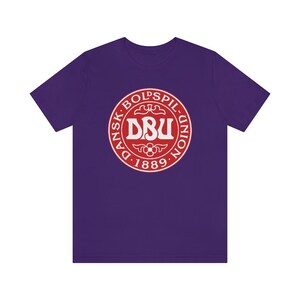 Puede incluir: Camiseta morada con un logotipo rojo y blanco que dice "Dansk Boldspil-Union D. 1889."
