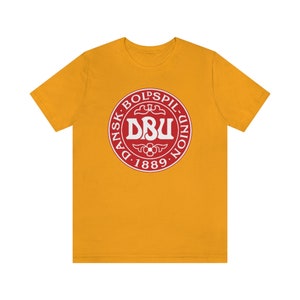 Puede incluir: Una camiseta amarilla con un logotipo rojo y blanco que dice "Dansk Boldspil-Union DBU 1889".