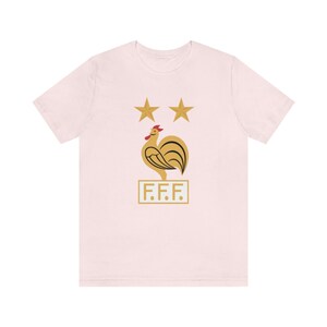 May include: A pink t-shirt with a gold rooster and two stars above it. The rooster is standing on a white rectangle with the letters "F.F.F." printed on it.