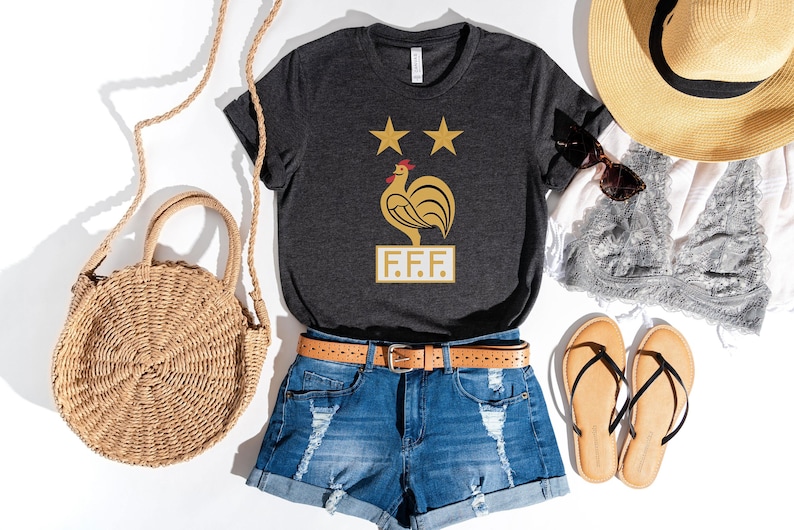 May include: A dark gray t-shirt with a gold rooster and two stars, the text "F.F.F." below the rooster. The shirt is paired with denim shorts, a brown belt, a straw hat, a straw bag, and sandals.