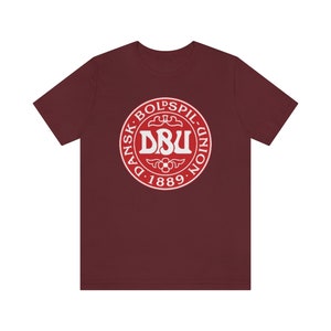 Puede incluir: Una camiseta de color granate con un logotipo circular blanco. El logotipo presenta el texto "Dansk Boldspil-Union D. 1889" y las letras "DBU" en una fuente m&aacute;s grande.