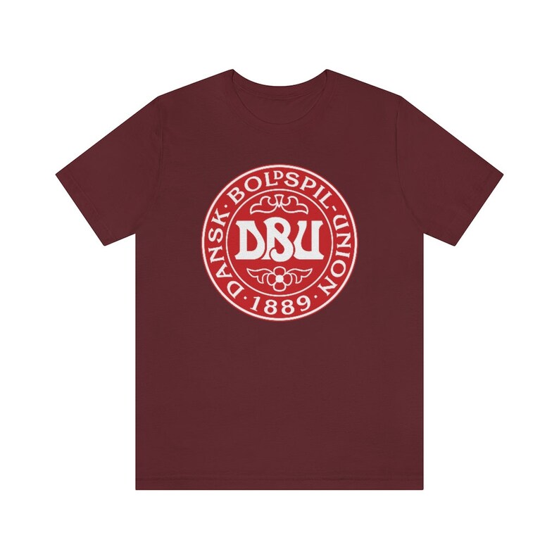 Puede incluir: Una camiseta de color granate con un logotipo circular blanco. El logotipo presenta el texto "Dansk Boldspil-Union D. 1889" y las letras "DBU" en una fuente m&aacute;s grande.