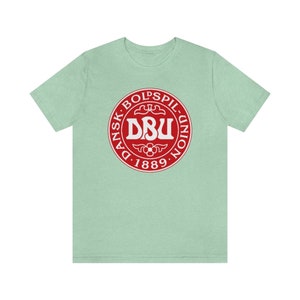 Puede incluir: Una camiseta verde claro con un logotipo circular rojo. El logotipo dice "Dansk Boldspil-Union DBU 1889" en texto blanco.