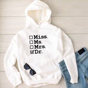 May include: White hooded sweatshirt with a black graphic that says "Miss. Ms. Mrs. Dr." with a tick next to "Dr."