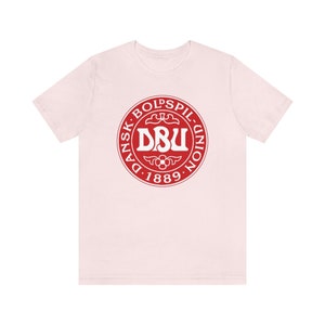 Puede incluir: Una camiseta rosa con un logotipo circular rojo. El logotipo dice "Dansk Boldspil-Union DBU 1889" en letras blancas.