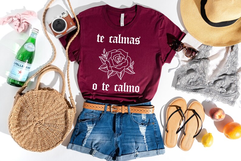 May include: A maroon t-shirt with a white rose and the text "te calmas o te calmo". The shirt is laid out on a white surface with a straw bag, a pair of sandals, a straw hat, a bottle of water, a camera, and a pair of sunglasses.