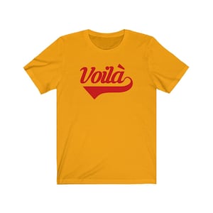 Puede incluir: Una camiseta amarilla de manga corta con un gr&aacute;fico rojo "Voila!" en una fuente retro.