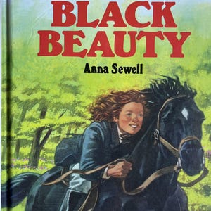 Op de afbeelding: Vintage boekomslag voor "Black Beauty" van Anna Sewell. De omslag toont een jong persoon die op een zwart paard rijdt door een groen bosgebied. De titel staat in grote rode letters.