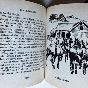 Op de afbeelding: Een open boek met tekst en een zwart-wit illustratie. De illustratie toont een nieuwe stal met verschillende paarden en twee personen. De tekst op de pagina luidt "BLACK BEAUTY" en "A New Stable."