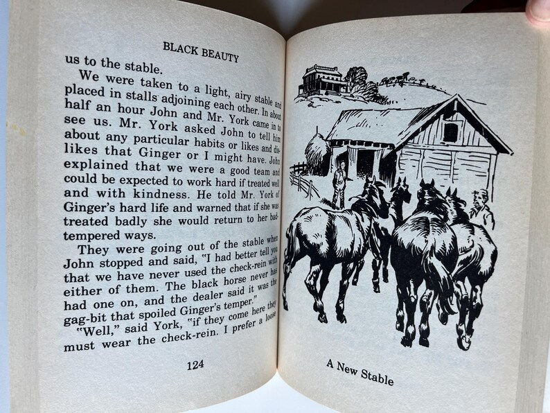 Op de afbeelding: Een open boek met tekst en een zwart-wit illustratie. De illustratie toont een nieuwe stal met verschillende paarden en twee personen. De tekst op de pagina luidt "BLACK BEAUTY" en "A New Stable."