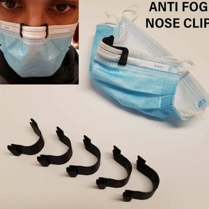 Op de afbeelding: Vijf zwarte plastic anti-condens neusclips voor gezichtsmaskers. De clips zijn U-vormig en hebben aan elk uiteinde een klein lipje. De tekst "ANTI FOG NOSE CLIP" is in wit op een blauw gezichtsmasker gedrukt.