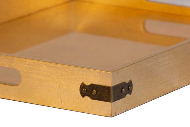May include: A gold-colored wooden tray with a clear glass insert and brass corner accents. The tray has a rectangular shape and is perfect for serving snacks or drinks.