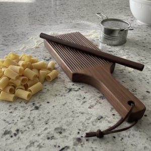 May include: A wooden gnocchi board with a dark brown finish and a matching rolling pin. The board has parallel grooves for shaping pasta. A pile of rigatoni pasta is to the left. A small metal sieve and a bowl are in the background.
