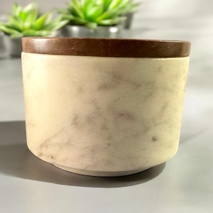 Puede incluir: Un recipiente cilíndrico de mármol blanquecino con un borde marrón oscuro. El recipiente tiene una superficie ligeramente texturizada con sutiles vetas grises. El diseño es simple y elegante, adecuado para diversos usos.