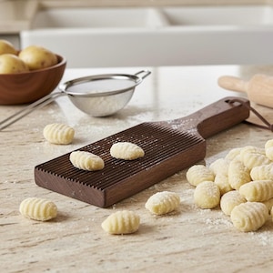 May include: A wooden gnocchi board with a grooved surface, alongside freshly made gnocchi. The board is brown, and the gnocchi are light yellow, dusted with flour. A bowl of potatoes and a sieve are in the background, suggesting a homemade pasta preparation.