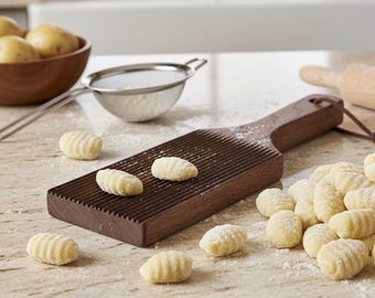 May include: A wooden gnocchi board with a grooved surface, alongside freshly made gnocchi. The board is brown, and the gnocchi are light yellow, dusted with flour. A bowl of potatoes and a sieve are in the background, suggesting a homemade pasta preparation.
