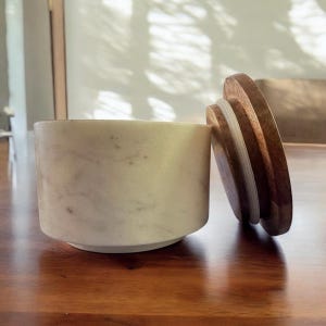May include: A cylindrical marble container with a wooden lid. The container is off-white with subtle gray veining. The lid is a warm brown, partially covering the opening. The design is simple and elegant.