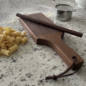 May include: A dark brown wooden gnocchi board with a ridged surface and a matching wooden rolling pin. The board features a handle with a hole and a brown leather cord attached. A small metal sifter and a pile of dry pasta are nearby.
