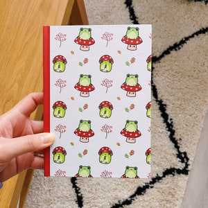 Peut inclure: Un carnet blanc avec une reliure rouge présente un motif répété de grenouilles vertes assises sur des champignons rouges et blancs. Les grenouilles sourient toutes et ont des yeux noirs. Les champignons ont des points blancs.