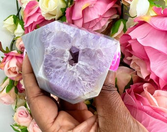 May include: A large, faceted amethyst geode crystal with a light purple interior and a gray and white exterior. The geode is held in a hand and surrounded by pink and white roses, creating a striking contrast.