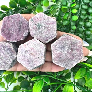 May include: Five pink ruby crystals in a hexagonal shape, with a rough, natural texture. The crystals are arranged in a hand, with the tips of the crystals pointing towards the viewer.