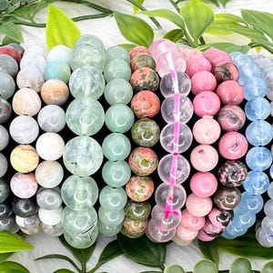 May include: A set of ten bracelets made with various semi-precious stones. The bracelets are made with different colors of stones, including green, pink, blue, and clear. The bracelets are stacked on top of each other.