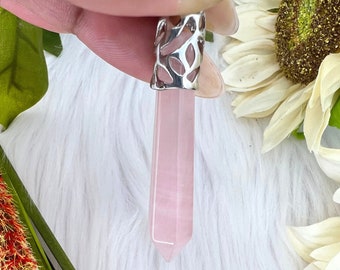 May include: A light pink rose quartz crystal pendant with a silver-colored metal cap. The crystal is a long, hexagonal shape. The pendant is held up by a hand, with a white and yellow flower in the background.
