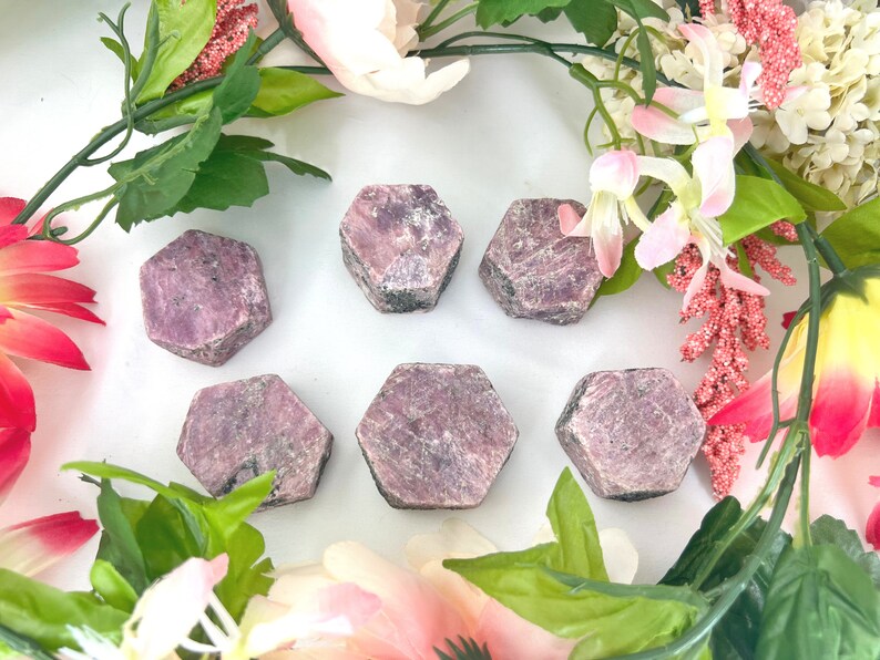 May include: Six pink ruby gemstone points, each with a hexagonal shape, are arranged on a white surface. The gemstones are surrounded by pink and white flowers.
