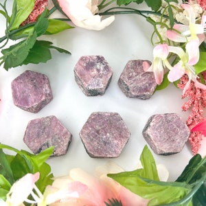 May include: Six pink ruby gemstone points, each with a hexagonal shape, are arranged on a white surface. The gemstones are surrounded by pink and green flowers.
