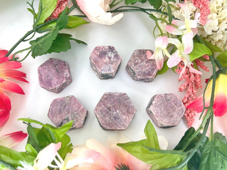 May include: Six pink ruby gemstone points, each with a hexagonal shape, are arranged on a white surface. The gemstones are surrounded by pink and green flowers.