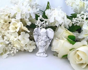 May include: A white and gray marble angel figurine with outstretched wings, set against a backdrop of white flowers and green leaves. The angel has detailed carving and stands upright. The flowers include roses and other varieties.
