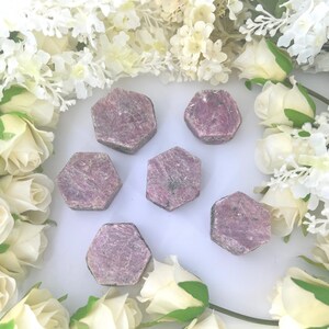 May include: Six pink ruby gemstone points in a hexagonal shape, arranged on a white surface. The gemstones are surrounded by white flowers.