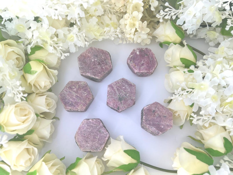 May include: Six pink ruby gemstone points in a hexagonal shape, arranged on a white surface. The gemstones are surrounded by white flowers.