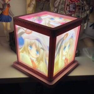 May include: A square, pink-framed light box featuring illuminated panels on the sides and top. Each panel displays a graphic illustration of a blonde character with large blue eyes wearing a white hat with a circular orange accent.