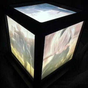 Può includere: Una scatola d'arte quadrata e illuminata con una cornice scura. I lati mostrano un'immagine morbida ed eterea di una persona con capelli fluenti e un paesaggio. Il pannello superiore mostra un cielo nuvoloso. La scatola emette un bagliore morbido e caldo.