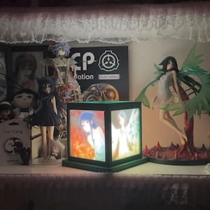 以下が含まれることがあります： 棚には、さまざまなアニメをテーマにしたフィギュアやアートワークが飾られています。 アニメのアートが描かれた、光る立方体型のランプが中央にあります。 他のアイテムには、額入りの絵や「SCP Foundation」と書かれた看板などがあります。
