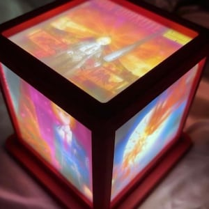 May include: A red, cube-shaped lamp with illuminated panels displaying colorful artwork. The lamp has a dark red frame and base. The artwork on the panels features a variety of colors, including orange, pink, and blue.