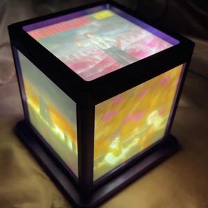 May include: A black, cube-shaped lamp with illuminated panels displaying a colourful image. The top panel shows text and a figure. The side panels feature a landscape with pink and yellow hues. The lamp sits on a square base.