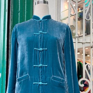 Può includere: Un cappotto di velluto color teal con colletto alla coreana e chiusure a frog. Il cappotto a maniche lunghe presenta due tasche anteriori ed è esposto su un manichino. La lunghezza del cappotto è di circa 102 cm.