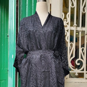 May include: A black patterned robe with long sleeves, a V-neck, and a matching belt. The robe is displayed on a mannequin. The background features a green wall and a white door with decorative ironwork.